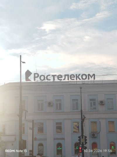 The image shows a bright, slightly hazy day in Yakutsk, Russia. The main subject is a building with a prominent sign that reads "Ростелеком" (Rostelecom) in large, dark letters. The building itself is light blue with white trim and multiple windows. The sky is a pale blue with scattered white clouds, suggesting either early morning or late afternoon.

In the foreground, a lamppost stands to the left, and a traffic light with red and green signals is visible on the right. The lower part of the image shows the street level, with glimpses of people and storefronts, including signs that say "КАНЦМИР" (Kantsmir) and "КРЕАТИВ" (Kreativ), and "КНИГИ" (Knigi). The overall atmosphere is calm and urban.

The time stamp "30.06.2026 19:56" is displayed in the bottom right corner, indicating it is late evening on June 30, 2026. The "REDMI Note 13" watermark appears on the bottom left. The light suggests it might be near sunset, casting a soft glow on the scene.
