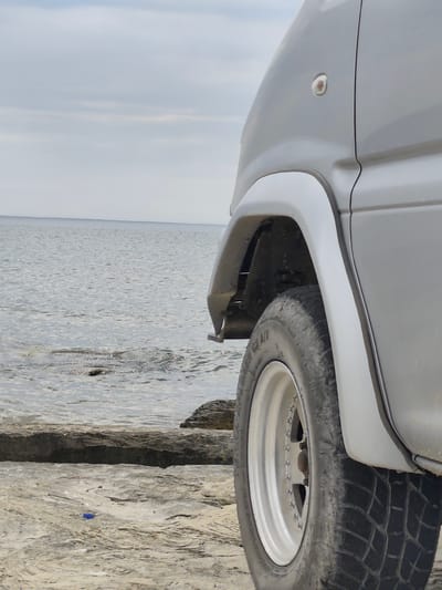 This is a medium shot, eye-level, outdoor photograph of a silver van parked on a rocky shore by the Caspian Sea in Aqshuqyr, Kazakhstan. The van is positioned in the right half of the frame, with its front wheel and fender visible. The wheel is a polished alloy rim with a black tire. The van's bodywork is a smooth, light gray color, and a small amber turn signal is visible on the side. The ground beneath the van is a mixture of sand and rough, layered rocks. In the background, the calm, grey waters of the Caspian Sea extend to the horizon, meeting a pale, overcast sky. The water is slightly choppy in places where it meets the rocks. A few scattered rocks are visible in the water.