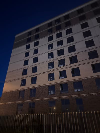 The image shows a tall apartment building with a dark blue sky in the background. The building has multiple floors, each with several dark windows. The lower floors are constructed with dark brick, while the upper floors are a lighter beige color. A fence runs along the bottom of the image, in front of the building. The building appears to be in a city setting, and based on the query, it is located in Petropavl, Kazakhstan. The time of day is likely evening or night, given the dark sky and the appearance of the windows. There are no people visible in the image.