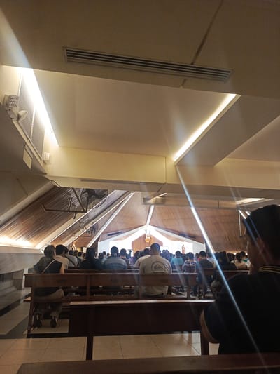 The image depicts the interior of a spacious building, likely a church or a large assembly hall, where a gathering is in progress. Numerous individuals are seated in neat rows of dark wooden pews, predominantly facing away from the camera towards the front of the hall.

The architecture features a high, multi-angled ceiling with sections of warm wooden paneling contrasted with lighter-colored beams and panels. Modern linear light fixtures are integrated into the ceiling, casting bright illumination and creating visible rays of light within the space. Overhead, air conditioning vents and large industrial-style fans are visible, indicating climate control.

At the distant front of the hall, a brightly lit white background or screen is discernible, with a figure, possibly a statue or a person, dressed in reddish-orange clothing standing before it. The people in the pews appear to be attentively observing the front. In the foreground, the back of a man's head and shoulders are visible on the right, also seated in a pew. Another man further down the aisle is wearing a grey t-shirt with the brand name "QUIKSILVER" visible on its back. The floor is covered with light-colored tiles. The scene conveys a sense of quiet observance or participation in a structured event, set in Guayaquil, Ecuador.