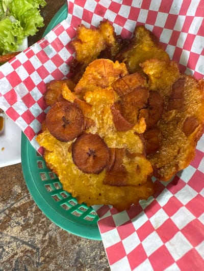 The image displays a close-up, top-down view of fried plantains or bananas served in a green plastic basket lined with red and white checkered paper. The dish consists of both flatter, golden-yellow pieces resembling tostones (fried green plantains) and several circular, darker brown slices that appear to be tajadas (fried ripe plantains). Some pieces exhibit a slightly irregular, crinkled texture from frying. In the upper-left background, out of focus, a small portion of fresh green lettuce is visible on a white surface, suggesting part of a meal. The item rests on a dark, mottled brown and grey table surface. The specific location in La Ceiba, Honduras, cannot be determined from the visual evidence provided. No people, interactions, or discernible actions are present in the frame.