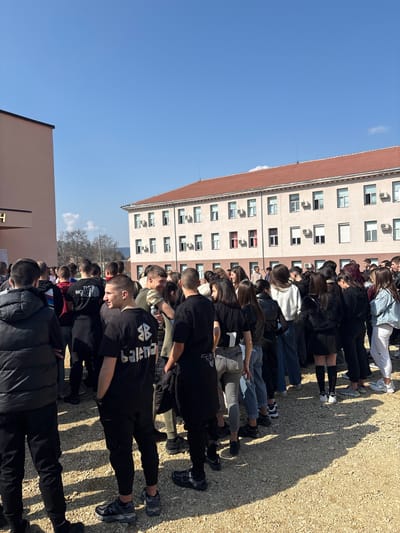 The image displays an outdoor gathering of approximately 50-70 individuals, predominantly young adults or teenagers, located in Cherven Bryag, Bulgaria. The scene is illuminated by direct sunlight under a clear blue sky, casting defined shadows on the light-colored gravel ground. In the background, a prominent four-story building with a light beige facade and a reddish-brown tiled roof is visible. This building features multiple rectangular windows, many equipped with external air conditioning units, and some with visible red interior blinds or shutters. To the far left, a section of a peach-colored building with a dark roofline is partially visible. Between the buildings, bare deciduous trees are present.

The gathered individuals are standing in a dense group, dressed in casual clothing. Predominant attire includes t-shirts, hoodies, jackets, jeans, and sneakers, with a frequent use of black and other dark colors. Notable clothing items include a black t-shirt with "THE NORTH FACE" text, a black t-shirt displaying a "BB balenciaga" logo, and an olive green camouflage t-shirt worn by a male holding a water bottle. A female is observed wearing a light blue denim jacket over white clothing. The individuals appear to be standing in close proximity, suggesting a social gathering or a period of waiting. No overt organized activity is discernible.