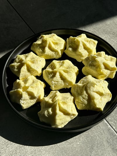 The image displays eight pale yellow, irregularly shaped solid pieces, possibly meringues, cookies, or another type of food, arranged on a round, matte black plate. Each piece exhibits a textured, somewhat crumbly or porous surface, suggesting a delicate or sugary consistency. They are scattered rather than neatly arranged on the plate.

The scene is illuminated by strong, directional natural light, likely sunlight, casting sharp, elongated shadows onto the plate and the surrounding surface. These shadows create a high-contrast visual, highlighting the contours of the objects and the rim of the plate.

The background features a light grey tiled surface, indicating a floor or countertop, with clear grout lines forming a grid pattern. Part of the tiled area is brightly lit by the sunlight, while another significant portion is in deep shadow, emphasizing the strong light source. The overall aesthetic is minimalist, focusing on the texture and form of the pieces and the interplay of light and shadow. There is no visible text in the image. The setting is an indoor environment, likely during the daytime, in Tomilino, Russia.