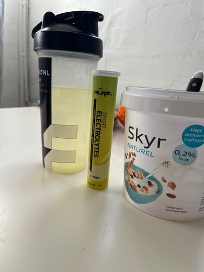The scene depicts an indoor setting, an unidentifiable room within Odense, Denmark. A white, textured wall forms the background, while the foreground is a flat, light-colored surface, likely a table or countertop. No individuals are present.

Three primary items are positioned on the surface:
1.  A transparent plastic shaker bottle with a black lid, positioned on the left. It contains a pale yellow liquid, filling approximately 30-40% of the bottle's volume. Partial branding "CTNL" is visible on its side.
2.  A cylindrical, yellow-green effervescent tablet tube, positioned centrally. The label reads "murph. SPORT ELECTROLYTES", with "CITRUS" flavor and "20 TABS • 78G". "VITAMINS • MAGNESIUM • SODIUM" is also visible.
3.  A large, white plastic tub of Skyr, positioned on the right. The label displays "Skyr NATUREL", "Højt protein indhold" (High protein content), and "0,2% fedt" (0.2% fat). A black-handled spoon is partially inserted into the tub's contents.