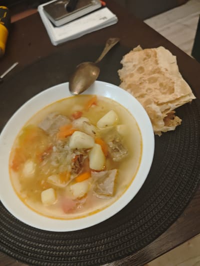 A close-up shot shows a plate of soup, a spoon, and a stack of flatbreads on a dark brown woven placemat. The soup, served in a white bowl, contains chunks of meat, potatoes, carrots, and cabbage in a light broth. An antique spoon rests on the edge of the bowl. A pile of folded flatbreads sits next to the soup. The scene appears to be a meal setting indoors, potentially a home. A notebook and a tool lie out of focus in the background, suggesting a cluttered table. No information indicates the location is in the city of Свистуха (кузяевский С/о), Russia.