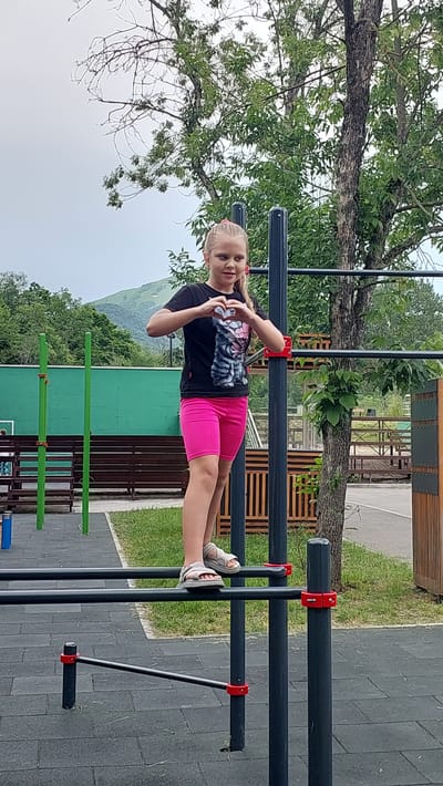 A young girl with blonde hair tied in pigtails is performing a planche on a set of parallel bars in an outdoor gym. She is wearing a black t-shirt with a cat graphic and bright pink shorts. Her feet are resting on the lower bar, and her body is extended horizontally. The gym equipment is made of dark metal bars with red connectors. The background features a large tree with green leaves, a grassy area, and a mountainous landscape under a cloudy sky. In the distance, there are wooden structures and other pieces of gym equipment. The scene appears to be set in the daytime, possibly late morning or afternoon, with overcast weather. The girl's facial expression suggests concentration and effort. The location is identified as Lermontov, Russia, which is known for its mountainous scenery.