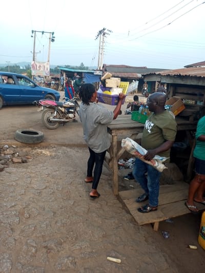 A woman in a grey shirt and black leggings argues with a man in a green shirt holding a bag of rice at an outdoor market. The woman is holding a basket of goods over her head. A blue car and a motorcycle are visible in the background. The market appears to be in a rural area, with buildings and power lines in the distance. An old tire lies on the rocky ground.
