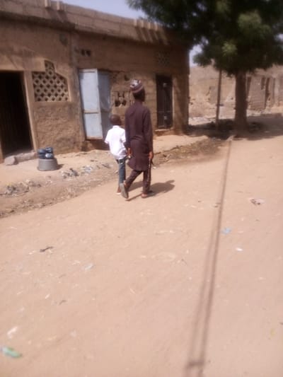 The scene depicts an unpaved dirt path in Rangaza, Nigeria, under a clear, bright sky. Two individuals walk away from the camera along the path. The taller individual, positioned right of center, wears a dark brown long tunic, dark trousers, and a patterned cap, carrying a stick or staff-like object in their right hand. To their immediate left and slightly behind, a shorter individual wears a white short-sleeved shirt and blue jeans.
On the left side of the image, a single-story structure with an earthen-toned, rough-textured facade is visible. It features a dark doorway on the far left, a window with a lattice pattern, a light-colored metal door that appears partially ajar, and another dark door further right. A grey metal basin containing several dark blue or black plastic bags rests near the base of the building, to the left of the lattice window. Scattered debris, including small plastic and paper fragments, litters the path and the area near the building.
To the right of the walking figures, a medium-sized tree with green foliage and a visible trunk stands. In the far background, another earthen-toned structure or wall is visible. Long shadows are cast on the ground, notably a distinct, thin, dark shadow extending across the foreground from the right side of the frame.