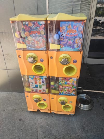 The image depicts a stack of four orange and yellow toy vending machines positioned on a grey, paved surface in front of a building. The building features a textured, grey wall and large glass doors with a dark frame, suggesting a commercial establishment. To the right of the vending machines, a metallic pet food or water bowl rests on the pavement. The vending machines contain various colorful toys and display cards, including "Jumbo Ocean," "Moshy Dangling," "Bubble Tea," and "Jumbo Festival Series." The machines have coin slots and dispensing chutes. The overall environment suggests an outdoor, public area, likely in an urban setting. The specific location of Vitoria-Gasteiz, Spain, is not discernible from the visual information alone, as there are no identifying signs or landmarks present in the image.