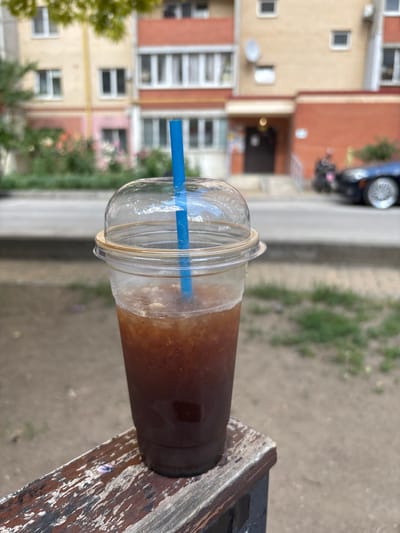 A close-up shot captures an iced coffee in a clear plastic cup with a domed lid and a blue straw. The beverage is dark brown with ice visible throughout. The cup is positioned on a weathered wooden surface in the foreground, which appears to be part of a bench or railing. In the background, a building with a light yellow and orange facade is visible, featuring multiple windows and balconies. The scene is set outdoors, with greenery and a street with a car and a motorcycle also present. The overall impression is of a casual, urban setting, possibly in Lymanka, Ukraine, given the architectural style and the presence of the vehicle.