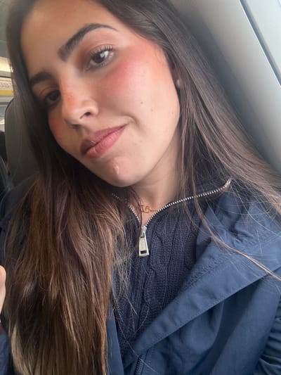 A young woman with long brown hair is pictured from a close-up, slightly angled perspective. She is wearing a dark blue jacket with a visible "POLO" zipper pull. Her makeup includes blush and lip color. The setting suggests she is indoors, possibly near a window, but the exact location within San Antonio, United States of America, cannot be determined from the provided image.