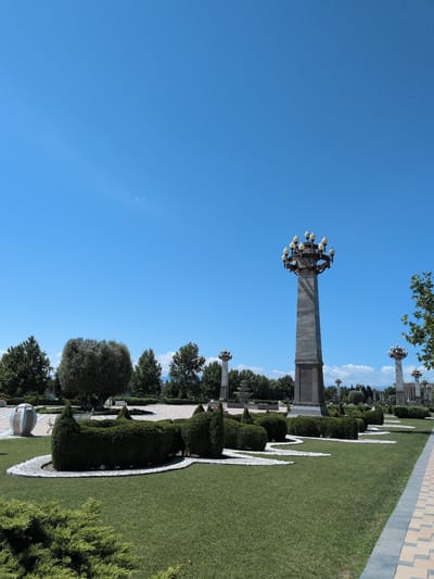 The image depicts a park area in Ganja City, Azerbaijan, under a clear blue sky. The foreground is dominated by a well-maintained green lawn with several elaborately shaped hedges. White gravel pathways wind through the manicured shrubbery, creating a decorative pattern.
In the mid-ground, several tall, ornate lampposts stand prominently. The most striking is a large, grey obelisk-shaped lamppost with a cluster of lights at its apex, featuring a decorative base. To the left of this, a modern, oval-shaped trash receptacle is visible. Further into the park, other lampposts and decorative elements are present, along with more trees and shrubs.
The background shows a line of trees, some appearing mature and lush. In the distance, faint clouds are visible against the sky, and distant structures or landforms can be perceived. The overall scene suggests a public park or garden designed for leisure and aesthetic appeal. No people are clearly visible in the image.