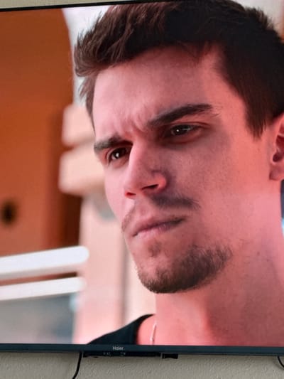 The image shows a close-up of a young man's face. He has dark hair, dark eyebrows, and a faint mustache and beard. His eyes are looking to his left, and his expression appears serious or contemplative. The lighting suggests an indoor setting with warm tones. The background is blurred, but appears to be some sort of structure with light-colored elements. The bottom of a television screen with the "Haier" logo is visible at the very bottom of the image, indicating the man's face is likely a still from a video being displayed on the screen. There is no discernible information about the city of Uyutnoye, Ukraine, within the image itself.