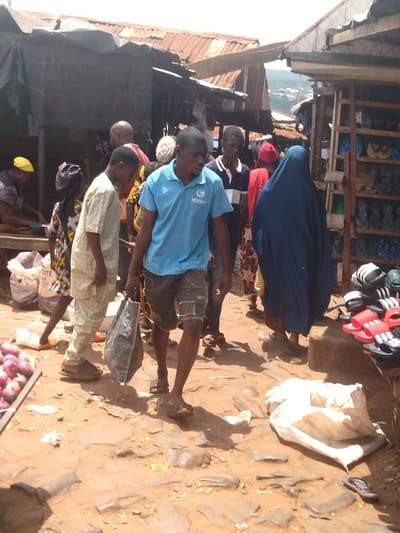 The image shows a bustling street scene in Lokoja, Nigeria, located at the coordinates 7.404 latitude and 7.632 longitude. It depicts a group of people walking on a dirt path lined with small, corrugated metal buildings. The path is uneven and covered in loose stones and debris. A man wearing a blue shirt and shorts is walking towards the camera, carrying a large black bag. He is surrounded by other pedestrians, including several women wearing traditional clothing. On the right side of the frame, a woman wearing a long blue robe walks past a stall displaying a variety of sandals. There are also bags of white grains on the ground, suggesting that this may be a market area. The image captures a snapshot of everyday life in this busy city.