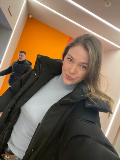 A woman poses for a selfie, slightly angled. She wears a black puffer jacket over a light blue turtleneck. Behind her and slightly below is a man also in a dark jacket. The background consists of a bright orange wall and a white ceiling with linear lighting fixtures. A smoke detector is visible on the ceiling. The scene appears to be inside a modern building.