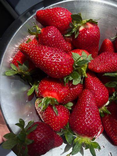 A close-up, high-angle shot depicts a collection of fresh strawberries in a metal bowl. The strawberries are a vibrant red, with tiny seeds and green leafy stems still attached. Water droplets are scattered across the strawberries and the surface of the bowl. The metal bowl has a brushed finish, reflecting light and creating highlights. The image appears to be taken in natural light. The location and city of Zovuni, Armenia, cannot be determined from the provided image.
