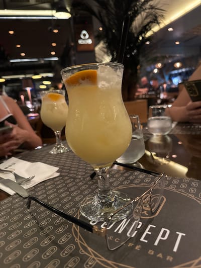 This image depicts an indoor evening scene at the Concept Café Bar, located in Strumica, North Macedonia. The primary focus is on a dark, reflective tabletop where a tall, clear cocktail glass filled with a light yellow, frothy drink, garnished with an orange slice and ice, stands prominently. Resting on a dark brown placemat/menu in front of this glass are a pair of dark-framed eyeglasses with clear lenses, displaying "ESCALADE CE" on the temple arm. The placemat/menu features a repeating pattern of a stylized oval and "C" symbol, with "CONCEPT & CAFÉ BAR" printed in large white letters towards the bottom right.
To the left of the main drink, a white folded napkin, a silver fork, and a silver knife are visible. Further back and to the left, a second, identical cocktail glass with the same drink and garnish is partially visible and out of focus. To the right of the main drink, a small, clear glass of water is present.
At least three individuals are present, visible through their arms and partial torsos. On the far left, a person's arm, wearing a white strap top, is holding a smartphone. On the far right, another person's arm is visible, also holding a dark-cased smartphone with a visible camera bump. Both individuals appear to be engaged with their phones.
The background is softly blurred, revealing a dimly lit, upscale environment. Overhead, various artificial light fixtures cast warm glows, creating bokeh effects. Dark, feathery foliage, likely artificial palm trees, are part of the decor, adding to the lounge-like ambiance. A glowing, abstract logo is visible in the upper left background. The overall scene suggests a relaxed social gathering in a contemporary café or bar setting during the evening.