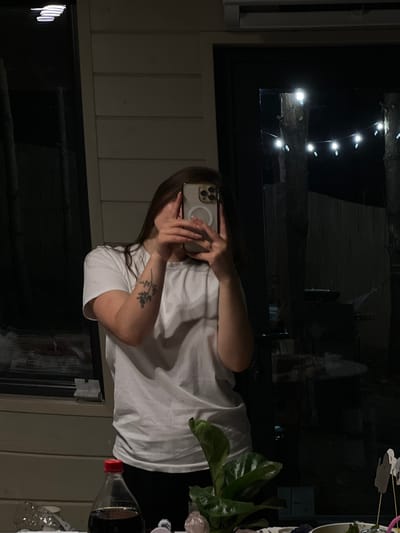 In Ivachiv Horishnii, Ukraine, a person is taking a mirror selfie outdoors. They are wearing a white t-shirt and black pants, and have a tattoo on their left arm. The person is holding a phone up to obscure their face. In the background, there is a glass door with string lights visible outside, and trees. A bottle of soda and a plant are on a table in the foreground.