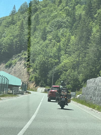 In the city of Lubnice, Montenegro, a red SUV is driving on a paved road lined with a forest of green trees on a steep hillside. A motorcycle with a rider wearing a helmet and a yellow vest is behind the SUV. The motorcycle is equipped with multiple storage boxes and a license plate that reads "MTO 56 80". On the left side of the road, there is a light blue building with a fence in front of it. The sky is clear and blue.
