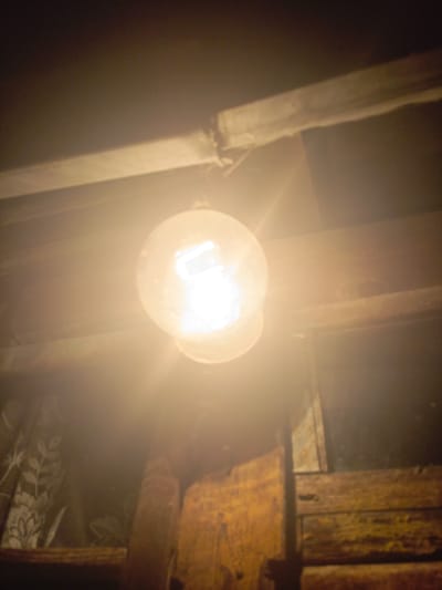 A dimly lit interior scene features a clear glass light bulb suspended from a wire, emitting a bright, warm light that illuminates the surrounding wooden beams and structures. The background is dark, with hints of patterned fabric on the left side. The overall atmosphere is rustic and somewhat aged. The location is identified as Ухманы, Russia. There are no people or animals visible in the frame.