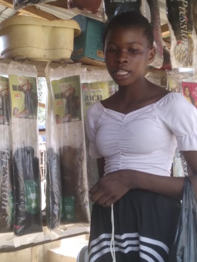 This image depicts a young Black female standing within a retail stall in Agege, Nigeria. The female subject, positioned slightly right of center, wears a white, ruched, short-sleeved crop top and a black skirt with white horizontal stripes near the hem. Her dark hair is short. Her mouth is slightly open, suggesting she may be speaking or in mid-sentence. Her left hand is visible, holding a black plastic bag.

The background reveals the commercial nature of the scene. To the left and behind the subject, multiple long, clear plastic packages of hair extensions are hanging vertically. These packages prominently display "X-Pression" and "RICH BRAID" branding, containing hair in various shades, including black and brown, with small green labels visible within the packaging. Above these, an overturned, light-colored plastic basin rests on a shelf. Behind the basin, stacked blue and brown cardboard boxes are present. To the right of the subject, more packaged hair extensions, some with "LUSCIOUS" branding, are hanging. The environment suggests an outdoor or semi-enclosed market stall, with daylight illuminating the scene.