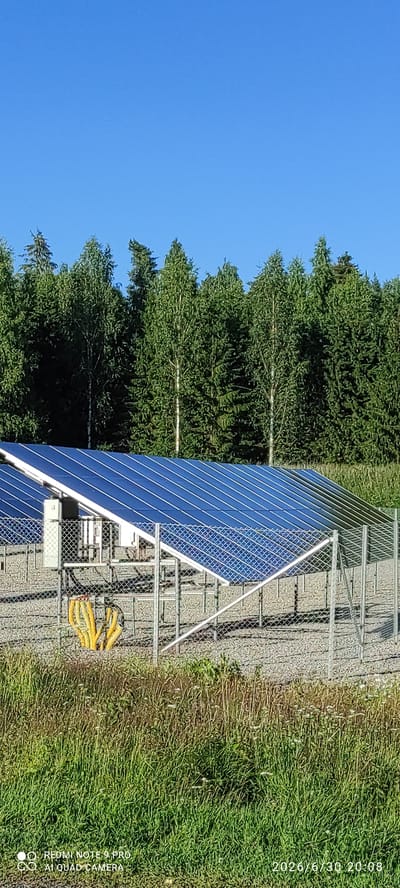 A solar panel farm is set against a backdrop of dense green trees under a clear blue sky. The solar panels are arranged in rows on a gravelly surface, supported by metal frames. A chain-link fence encloses the area, with yellow cables bundled together near the fence in the foreground. The overall scene is one of renewable energy infrastructure situated in a natural environment. The image includes a timestamp in the lower right corner, indicating the photo was taken on June 30, 2026, at 20:08. There are no people or animals visible in the image. The location is identified as Joutsa, Finland.