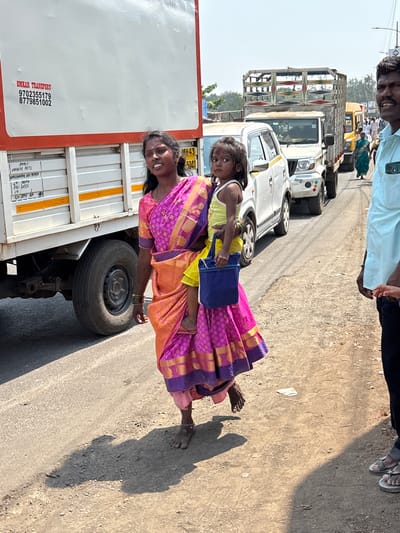 The image shows an Indian woman carrying a small child on her hip while standing in the street next to a parked truck. The woman is wearing a pink and orange sari. The child is wearing a yellow shirt and blue shorts and carrying a blue bag. The truck has the words "OMKAR TRANSPORT" and phone numbers "9702355179" and "8779851002" printed on its side. The license plate on the truck is "MH43 5041." In the background, there are other vehicles and people, suggesting a busy street. The text on the truck also mentions "KOPARKHAIRANE NAVI MUMBAI." It is not possible to verify the location in Ambernath, India, based solely on the image.