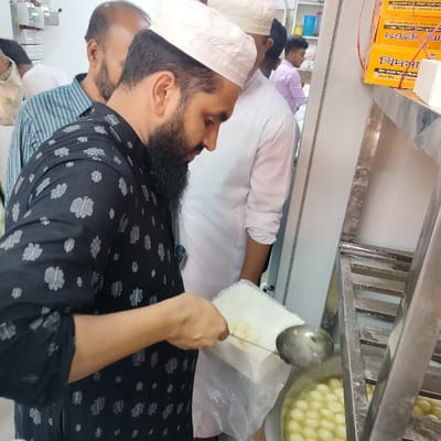 The scene takes place inside a shop located at 22.686 latitude and 90.645 longitude in the city of Bhola, Bangladesh. A man wearing a black shirt with a white floral pattern and a white skullcap is scooping a sweet, white, round dessert from a plastic container into a stainless steel pot filled with water. The dessert is in a rectangular, clear plastic container, and the man is using a metal ladle. Behind the man is another man wearing a white shirt and a white skullcap. The man in the white shirt is standing, and his right hand is on his hip. There is a third man in the background who is partially visible; he appears to be wearing a blue and green striped shirt. The shop's wall is painted white, and there are a few shelves along the wall to the right of the scene. There are yellow, red, and blue boxes on the shelves, but the writing on the boxes is illegible. The floor is light gray tile. The room is well-lit and clean.
