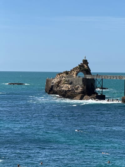 The image captures a sunny day at the Rocher de la Vierge (Rock of the Virgin) in Biarritz, France, a famous landmark situated on a rocky outcrop in the Atlantic Ocean. A narrow, ornate white footbridge, supported by dark metal beams, connects the mainland to the rock. Perched atop the highest point of the rock formation is a white statue of the Virgin Mary, a prominent feature of the site. The rough, textured surface of the rock is visible, with some areas appearing to be reinforced with stone structures. The sea is a vibrant blue, with gentle waves lapping at the base of the rocks and creating white foam. In the foreground, several individuals are swimming or floating in the ocean, enjoying the clear weather. The sky above is a bright, cloudless blue, typical of a summer day.