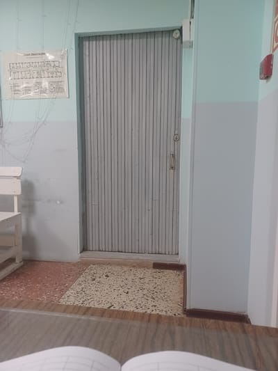 The image depicts an interior hallway with a light blue and grey painted wall, separated horizontally. On the left side of the wall, an evacuation plan is posted. Below the evacuation plan, a white wooden bench is visible. A large, grey metal rolling door dominates the center of the image. To the right of the door, a red fire alarm is mounted on the wall. The floor features a terrazzo pattern, with two distinct sections: one reddish-brown and the other light beige with grey speckles. In the foreground, a wooden surface, possibly a desk or table, is present, with the edge of an open notebook visible. The scene is devoid of people and any discernible activity. The location of Shudayag, Russia, is not visually ascertainable from the image itself.