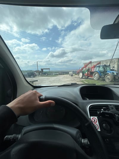 The image depicts a driver's perspective from inside a vehicle, looking out the front windshield in Srednia, Bulgaria. A right hand rests on a black, perforated leather steering wheel, with the dashboard visible below. Through the windshield, an unpaved lot or farmyard unfolds under a sky filled with scattered cumulus clouds. In the middle-ground to the right, a blue tractor is parked next to a light-colored concrete building, equipped with a large red boom sprayer attachment. Further left in the background, a green tractor, a dark SUV, and a white utility van are visible, along with various flatbed trailers and agricultural equipment. A small, dark animal (possibly a dog) stands near the white van. Utility poles and power lines traverse the upper half of the scene. Distant fields and hills are visible on the horizon beneath the cloudy sky.