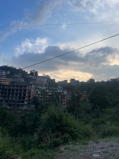 This is a landscape photograph of Dharamkot, India. The view is across a hillside densely packed with buildings. Many of the buildings are in various states of construction. The buildings are light-colored and contrast with the dark green vegetation. A bright, hazy sky sits above the cityscape. The hillside on the right is covered in dense foliage. A few utility wires are strung across the sky in the upper left.