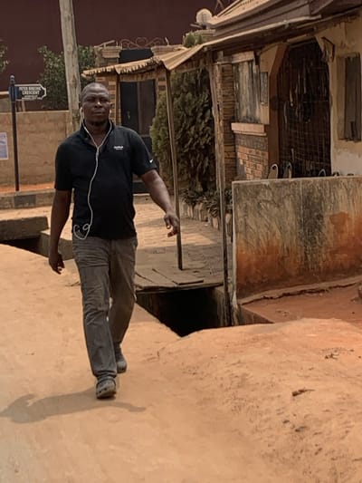 An adult male is captured walking on a reddish-brown dirt path in Otuocha, Nigeria. He is centrally positioned, moving forward and slightly to the right. The man wears a black short-sleeved polo shirt with a white logo on the left chest, grey trousers, and light-colored closed-toe shoes. White wired earphones are visible, with the wires extending from his ears down his torso. To the left, a street sign is visible, identifying "PETER OKECHE CRESCENT" and "MADONA SAFE LANE." An open concrete drainage ditch runs alongside the path on the right, partially bridged by wooden planks. Beyond the ditch to the right, a structure with a corrugated metal roof, exposed brickwork, and a barred opening is present. Green foliage and additional building structures are visible in the background. The scene is illuminated by daylight.