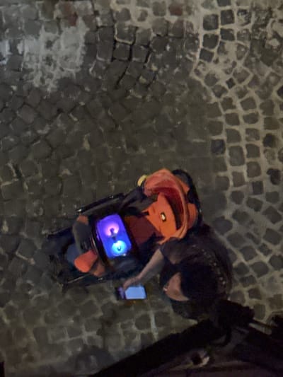 Observed from an elevated perspective on what appears to be a cobblestone street, potentially in Castelnuovo di Porto, Italy, the scene depicts an individual standing beside a brightly lit, compact object. The individual is partially visible, appearing to interact with a mobile phone in their hand. The brightly lit object is equipped with a screen displaying glowing, cartoonish eye-like features. The area surrounding the individual and the object is dimly lit, with the cobblestone pavement forming a significant portion of the visual background.