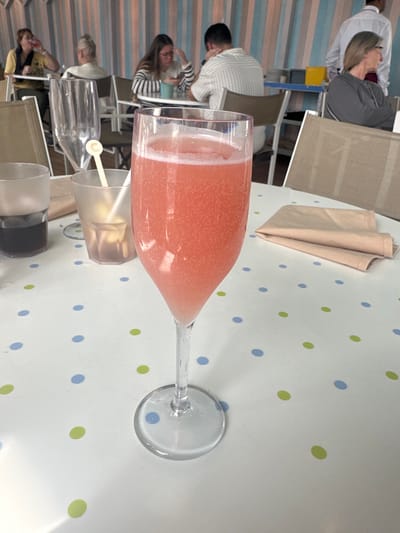 The scene depicts the interior of a casual dining establishment or cafe in Puerto del Rosario, Spain. In the immediate foreground, a clear, tall-stemmed glass containing a sparkling, light pink beverage with visible bubbles stands centered on a white tabletop. The tabletop features a scattered pattern of light blue and light green polka dots. To the left of this glass, a shorter, dark plastic cup holds a dark liquid, and a frosted plastic tumbler contains a light, clear liquid with a white plastic stirrer. A folded beige fabric napkin rests to the right of the central glass.

In the mid-ground and background, multiple individuals are seated at tables. On the left, a woman with dark hair wears a dark top and appears to be drinking from a glass. Behind her, another individual with light hair is partially visible. Further right, a woman with dark hair and glasses, wearing a horizontal striped shirt, is seated and looking down at a mobile phone. Across from her, a man with short dark hair, wearing a vertically striped shirt, is also seated and appears to be looking down. On the far right, a woman with light hair and glasses, dressed in a grey top, is seated and looking to her right. Behind her, a man in a white collared shirt, potentially a staff member, is standing. The background wall or partition displays vertical stripes in alternating shades of light blue, pinkish-beige, and white. The overall environment suggests a relaxed atmosphere typical of a cafe or restaurant.