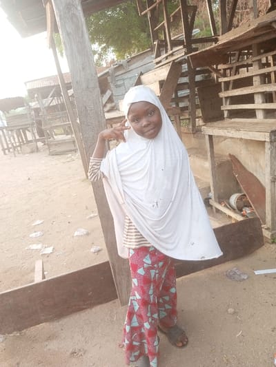 A young child, possibly a girl, with dark skin and a smiling expression, is the central subject of the portrait-oriented image. She wears a white hijab that covers her head and drapes over her shoulders and torso. Beneath the hijab, a horizontally striped long-sleeve shirt is visible. She also wears vibrant patterned trousers, primarily red with light blue and white geometric shapes, and dark sandals on her feet. Her left hand is raised, with her index and middle fingers extended in a "peace" sign or similar gesture, while her other fingers are curled.
The child is standing outdoors on what appears to be a sandy or dirt ground. The lighting suggests it is daytime, likely sunny, casting a bright glow on the scene. In the background, there are various wooden structures that look like makeshift stalls, unfinished buildings, or scaffolding. These structures are irregular and appear weathered, suggesting a rural or semi-urban setting. To the far left, a structure with a corrugated metal roof is visible. Green foliage can be seen in the distant background, hinting at vegetation. There is no visible text in the image. The child's pose and smile convey a sense of happiness and playfulness. The location context indicates this scene is in Ebiya, Nigeria.
