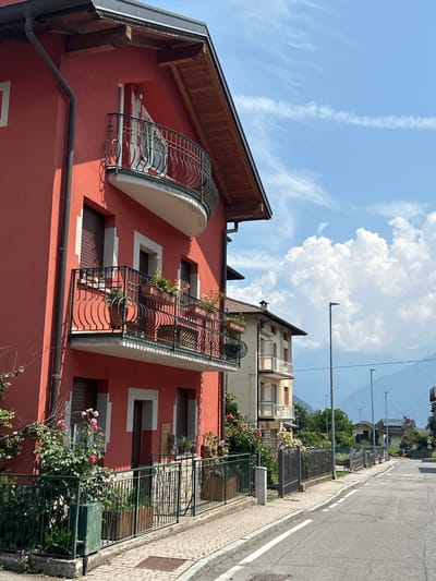 The image depicts a street scene in Ossimo Inferiore, Italy, on a sunny day. A vibrant red building with balconies featuring decorative wrought-iron railings dominates the left side of the frame. Several windows with closed shutters are visible on the building's facade, as are planters filled with flowers on the balconies. In front of the building, a green metal fence encloses a garden with rose bushes.

To the right of the red building, a lighter-colored building with balconies is visible. A paved sidewalk runs along the front of both buildings, adjacent to a two-lane asphalt road. The road curves gently to the right as it recedes into the distance, lined with streetlights and some houses. In the background, a range of hazy blue mountains can be seen under a bright blue sky with scattered white clouds. The overall atmosphere is tranquil and residential. No people are visible in the image.