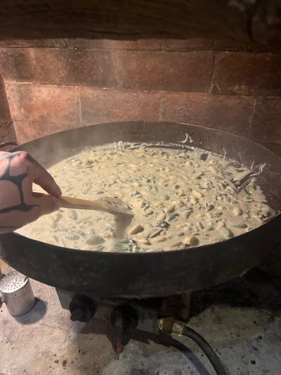 An overhead shot captures a person stirring a creamy pasta dish in a large, dark, round pan. The pan contains what appears to be gnocchi or similar ridged pasta shapes, submerged in a thick, light-colored sauce, with visible flecks of green (possibly herbs or vegetables). A wooden spoon is held by a hand, which is prominently tattooed with dark, intricate patterns covering the knuckles and back of the hand.
The cooking setup is rustic, suggesting an outdoor or traditional kitchen environment. To the left, a textured wall or pillar of reddish-brown brick or stone masonry forms the background. To the right, a rough, grey concrete or stone surface is visible, along with a black gas hose connected to a metallic fitting, indicating a gas burner or stove system below the pan. A metal component with multiple holes, possibly a part of the burner, is also visible near the bottom right.
The lighting is dim, casting shadows and highlighting the steam or vapor rising from the hot food, suggesting the scene takes place in the evening or at night. The focus is on the cooking process. No text is visible in the image. The activity takes place in Cordoba, Argentina.