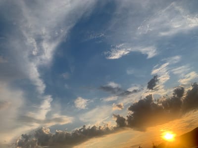 The image depicts a sky filled with a dynamic array of clouds during what appears to be either sunrise or sunset. The upper portion of the sky is a clear, deep blue, gradually transitioning towards lighter shades of blue and white near the horizon. Wispy, cirrus clouds are scattered throughout, some appearing feathery and delicate, while others are denser and more layered.
Towards the bottom and right of the frame, a vibrant sunset or sunrise is taking place. The sun is a bright, intense orb, casting a warm, golden-orange glow that illuminates the undersides of the clouds. These clouds are much darker, with shades of gray and brown, forming a dramatic contrast against the luminous sky. Some of these darker clouds appear thick and textured, suggesting they may be cumulonimbus or altocumulus formations catching the low-angled light.
The horizon line is not clearly defined due to the focus being on the sky, but there is a dark, silhouetted shape on the far right which could be a distant hill or mountainside. A few sparse tree branches or foliage are visible in the very bottom foreground, adding a subtle sense of scale.
The scene is devoid of any human presence or recognizable urban elements. Therefore, it is not possible to ascertain any specific location within the city of Khust, Ukraine, based solely on the visual information provided. The image captures a natural phenomenon and does not contain any contextual clues that would link it to a particular place or activity.