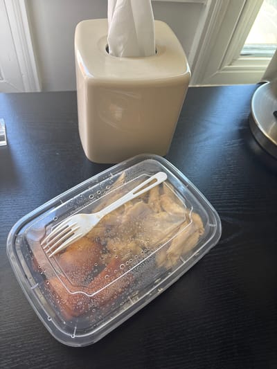 A close-up, high-angle shot shows a clear plastic food container with a white, compostable fork resting on top of the lid. The container is filled with what appears to be a meal of chicken and possibly rice. Condensation is visible on the inside of the lid. The container sits on a dark, textured surface, likely a table. In the background, a beige tissue box dispenses white tissues. The scene is illuminated by natural light coming from a window, casting soft shadows. The overall environment suggests an indoor setting, possibly a home or office, in San Francisco, United States of America.