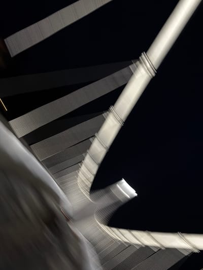 This media file displays an abstract night shot of a bridge structure. The dominant subjects are illuminated white and light grey structural elements, contrasting sharply against a dark, almost black background. These elements include a thick, main pylon or beam, extending diagonally across the frame, and numerous thinner, parallel cables or stays that radiate from it.
The pylon features distinct horizontal rings, while the cables display a ribbed or textured surface. Both the pylon and cables are rendered with significant motion blur, transforming them into luminous streaks and lines across the dark expanse. This effect suggests a long exposure photograph taken with camera movement or from a moving perspective.
No people or visible text are present. The image captures the bridge at night, illuminated by artificial light, creating a dynamic and almost ethereal representation of its architecture. Based on the provided context, the scene is a bridge in Osijek, Croatia, likely the Drava River bridge.