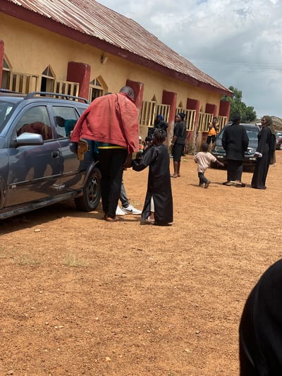 In Kassa, Nigeria, a group of people are gathered outside a building with a red corrugated metal roof and tan walls. A dark gray SUV is parked to the left, with its rear door open. A man in a red jacket and black pants stands near the car, looking at a young girl in a long black robe. The girl is facing away from the viewer. A toddler in a light-colored t-shirt and dark pants runs past them. Several other individuals, mostly women in dark clothing and headscarves, are visible in the background, some near another car. The ground is covered in reddish-brown dirt with scattered small stones and dry grass. The sky above is partly cloudy.