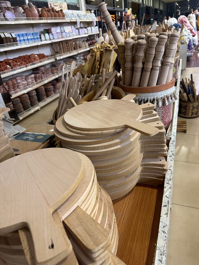 In Acıpayam, Turkey, a retail store displays numerous wooden kitchen utensils and serving boards. In the foreground, several stacks of round wooden serving boards with handles are arranged. To the right, a container holds an assortment of wooden rolling pins and other kitchen tools. In the background, shelves are stocked with various earthenware pots, packaged items, and more wooden utensils. The scene depicts a commercial environment focused on kitchenware.