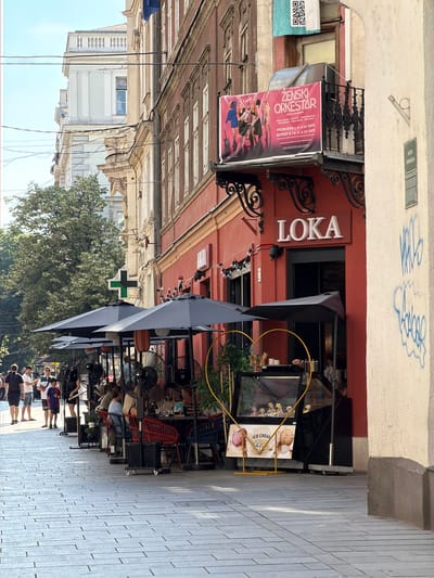 In Sarajevo, Bosnia and Herzegovina, a street-level outdoor seating area for a cafe or restaurant is captured on a sunny day. The foreground is a paved sidewalk made of light grey rectangular stones. To the right, a cream-colored building wall features prominent blue graffiti.
The main subject is a row of outdoor seating beneath dark grey umbrellas, occupied by several patrons. Fans are positioned around the seating area to provide cooling. In front of the establishment, a display case for ice cream is visible, adorned with a heart-shaped yellow frame. A sign in front of the display case advertises "PREMIUM ICE CREAM."
The building facade is painted a vibrant red and features an entrance with a sign that reads "LOKA" in white letters. Above the entrance, a balcony railing with decorative metalwork is present. A large banner advertising a "ŽENSKI ORKESTAR" (Women's Orchestra) is displayed on the building, along with a QR code.
Further down the street, trees and other buildings are visible. A green cross, typically signifying a pharmacy, is mounted on one of the buildings. Several people are walking along the sidewalk on the left side of the image, some appearing to be tourists. The overall atmosphere suggests a lively urban environment on a warm day.