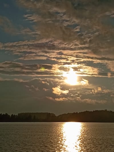 This is a horizontal, eye-level photograph of a lake and sky during sunset. The sun is low in the sky, partially obscured by clouds, casting a bright reflection on the water. The water is a muted brown-grey, with ripples catching the sunlight and creating a glittering effect. A dark, tree-lined silhouette of land stretches across the horizon, dividing the water and sky. The sky is a mix of blue, grey, and orange clouds, with the sun appearing as a bright white light in the center of the frame. The overall mood is serene and peaceful. The image captures a natural landscape, likely in Mołowiste, Poland, given the geographical context. There are no people or specific man-made objects visible in the scene, focusing solely on the natural elements of a sunset over water.