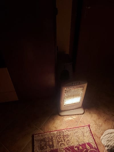 The image displays an indoor scene, likely at night or in a dimly lit room, with an electric heater as the central and brightest object. The heater, possibly a halogen or infrared model due to its glowing internal elements, has a light-colored casing with control buttons on its side. It rests on a textured, light brown tiled floor, casting a warm, orange-toned light that illuminates the surrounding area.
To the right of the heater, a rectangular rug with distinct purple/red and beige/white patterns is partially visible on the tiled floor. Further to the upper right, a white, soft-looking object, possibly a towel or laundry, is also partially in view. The left side of the frame is dominated by a dark, flat surface, likely a piece of furniture or a wall, which is largely in shadow, creating a strong contrast with the brightly lit heater and floor. No people are visible, and no specific activity other than the heater being operational is depicted, suggesting the space is being warmed. The general ambiance indicates a cozy or functional room setting.