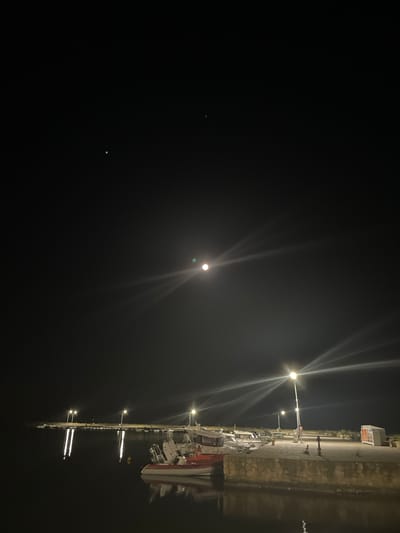 The image shows a nighttime scene at a harbor in Limenas Avdiron, Greece. The sky is dark with two visible celestial bodies, possibly planets or distant lights, one slightly greenish and the other brighter and white, with lens flares emanating from the latter. A dimly lit harbor area is visible in the lower half of the image. A concrete dock with stone foundations extends into the dark water. Several boats are moored along the dock, with one white and red boat prominently in the foreground. Lampposts illuminate the dock, casting reflections on the water. The atmosphere is calm and serene, suggesting a quiet night in the port.