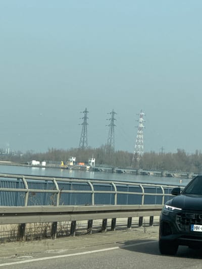 The image depicts a panoramic view from a moving vehicle on a bridge overlooking the Rhine River near Altenheim, Germany, under hazy conditions. In the immediate foreground, the front right section of a dark-colored SUV, bearing a German "HH-" license plate (Hamburg registration), is partially visible on the right. A gray metal guardrail and a blue metal barrier structure line the edge of the bridge. The midground features a wide body of water, likely the Rhine, with a dam or lock system visible across it, including multiple control buildings and industrial structures on the far bank. The distant background reveals a line of bare trees, above which three large lattice-type electricity transmission pylons are prominent. Two pylons are metallic grey, and the third (rightmost) is painted with alternating red and white bands. Further left in the background, industrial facilities with chimneys are faintly discernible. No individuals are present in the scene.