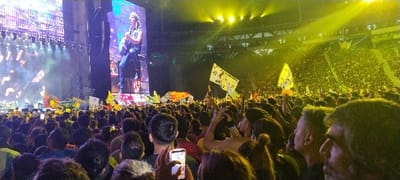 This image captures a dynamic indoor concert or live performance from the perspective of the audience in Tolosa, Argentina. A vast crowd fills the foreground and midground, mostly seen from behind, all facing a brilliantly lit stage. Many individuals are holding up mobile phones, capturing photos or videos of the event, with one phone screen clearly illuminated in the bottom-center.
The expansive stage features massive LED screens displaying a close-up of a male performer with long hair, possibly playing a guitar. The stage is illuminated with a blend of blue and purple lighting, while powerful yellow spotlights sweep across the upper tiers of the stadium, revealing more spectators filling the seats.
Throughout the standing crowd, several large yellow flags and banners are held high, adding to the energetic atmosphere. One such flag prominently displays the text "ETERNA PASION GRANDE". The event is clearly taking place at night, given the reliance on artificial lighting within the large stadium or arena. The scene conveys an enthusiastic and engaged mood among the concert-goers.