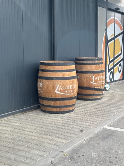 This media file shows two wooden barrels with black metal bands resting on a textured grey paved surface. The barrels are positioned side-by-side, with the top barrel slightly to the left and in front of the bottom barrel. Both barrels have white lettering on them. The top barrel features the text "3 АГРЕУС" and "ВИНАРНА". The bottom barrel has "7 AGREUS" and "WINERY" written on it. Behind the barrels is a dark grey, horizontally ribbed surface, possibly a garage door or industrial building facade. Above this, a portion of a window with a graphic design featuring orange and yellow elements is visible. The scene appears to be outdoors, with a grey sidewalk or road to the right of the barrels. The lighting suggests it is daytime. The setting seems to be in an urban or commercial area, possibly outside a winery or business. There are no people visible in the frame.